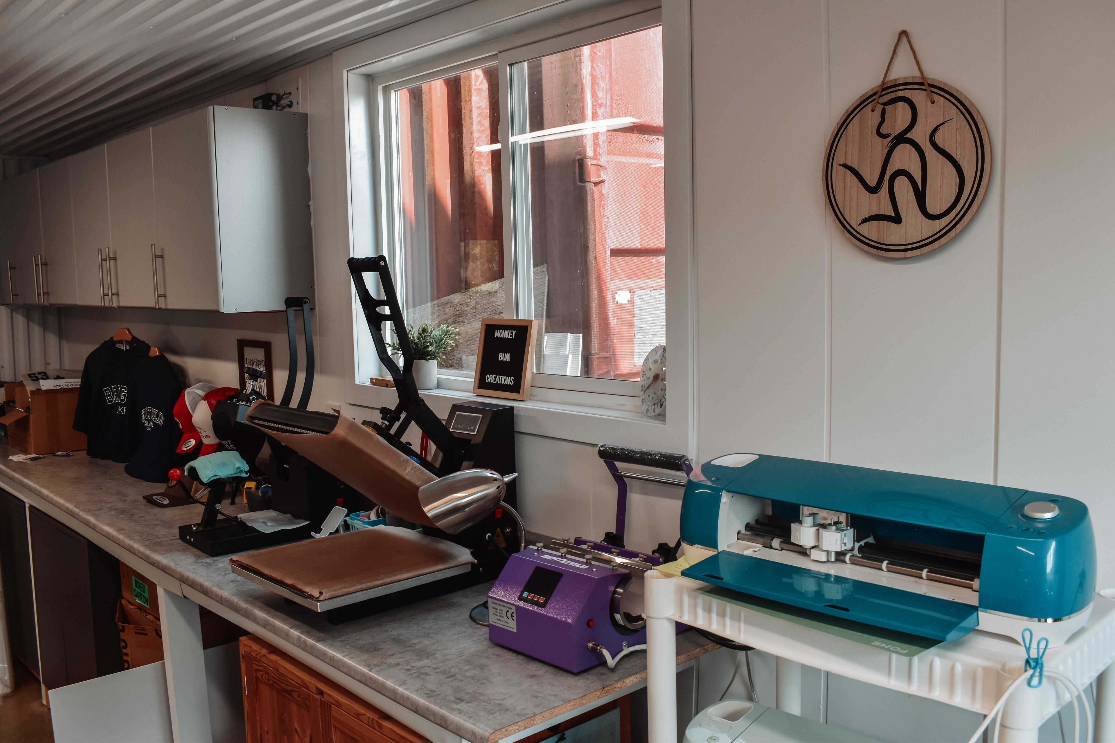Monkey Bum Creations workshop with a sewing machine, vinyl machine, heat press, desk, and various promotional products on a counter.